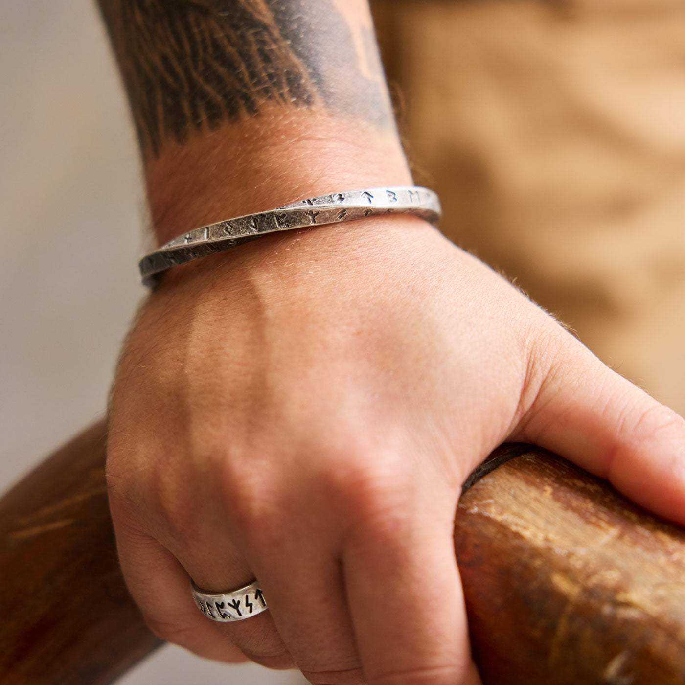 Close-up of a hand gripping a wooden railing, wearing a Viking-inspired stainless steel Rune Bracelet engraved with the Elder Futhark alphabet. A matching Rune Ring with the same ancient runes adorns the finger, symbolizing Norse heritage and mysticism.