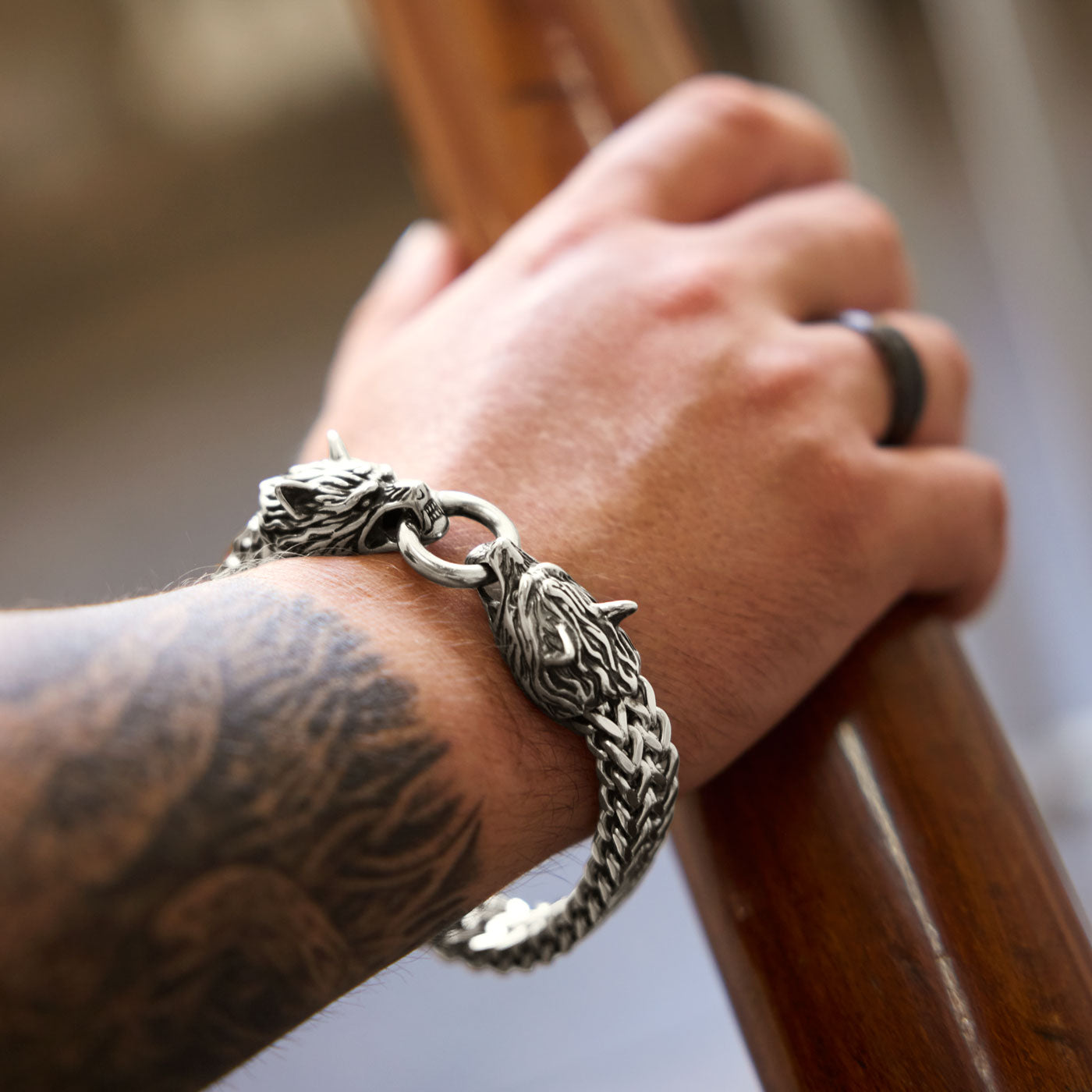 A stainless steel Geri and Freki wolf bracelet from Odin Trinkets worn on a tattooed wrist, featuring intricately crafted wolf heads biting onto a central ring, paired with an antique-style rune ring.