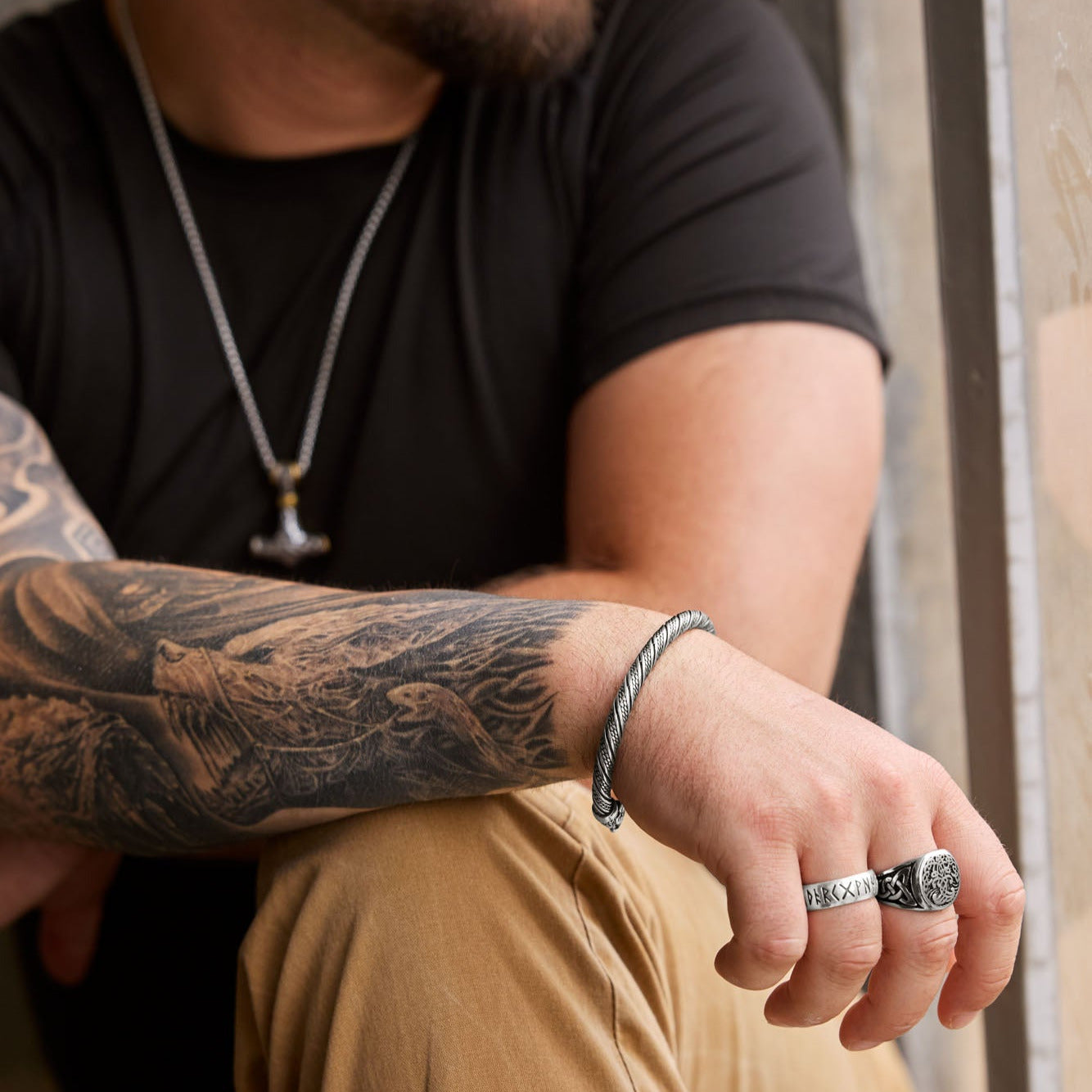 Man wearing the Mjölnir necklace with a Thor's Hammer pendant featuring intricate engravings and gold accents, paired with a twisted cuff bracelet, a silver rune ring showcasing the Elder Futhark alphabet, and a Yggdrasil ring symbolizing the Tree of Life, complemented by a tattooed arm and casual attire. Available at Odin Trinkets.