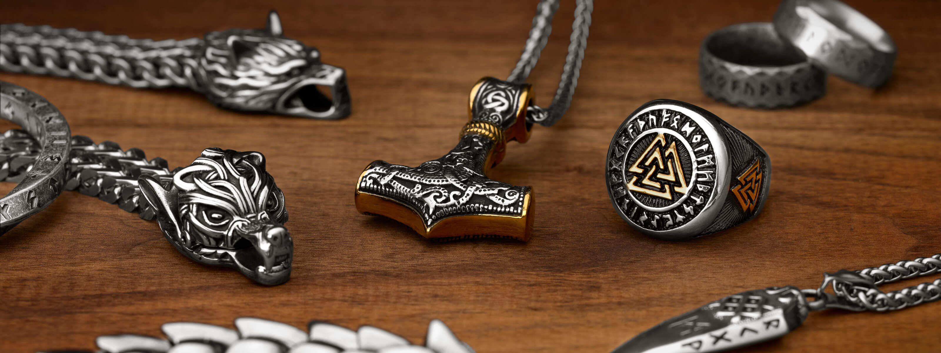 A Viking jewelry collection web banner displaying various bold pieces including a Mjölnir pendant, wolf head necklace, and a Viking ring featuring the Valknut symbol, arranged on a wooden surface.