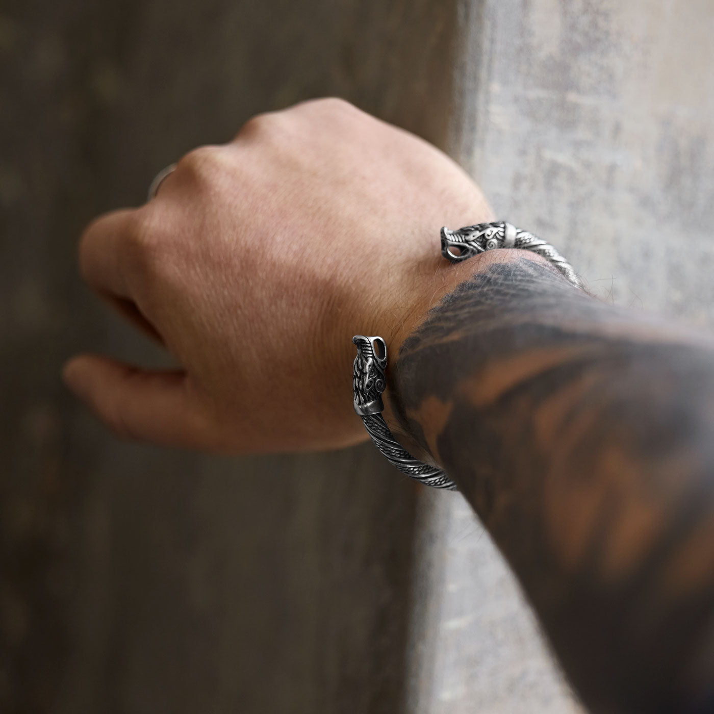 A Viking-style wolf bracelet worn on a wrist, featuring intricately designed wolf heads on both ends. The detailed bracelet coils around the wrist, displaying strong craftsmanship and a tattooed arm.