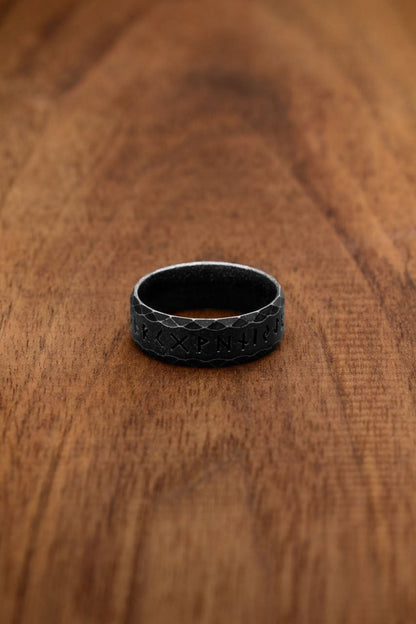 A black stainless steel rune ring, labeled as the "Antique" variation, featuring the engraved Elder Futhark alphabet. Its dark, matte finish and faceted texture create a bold and mysterious design inspired by Norse mythology.