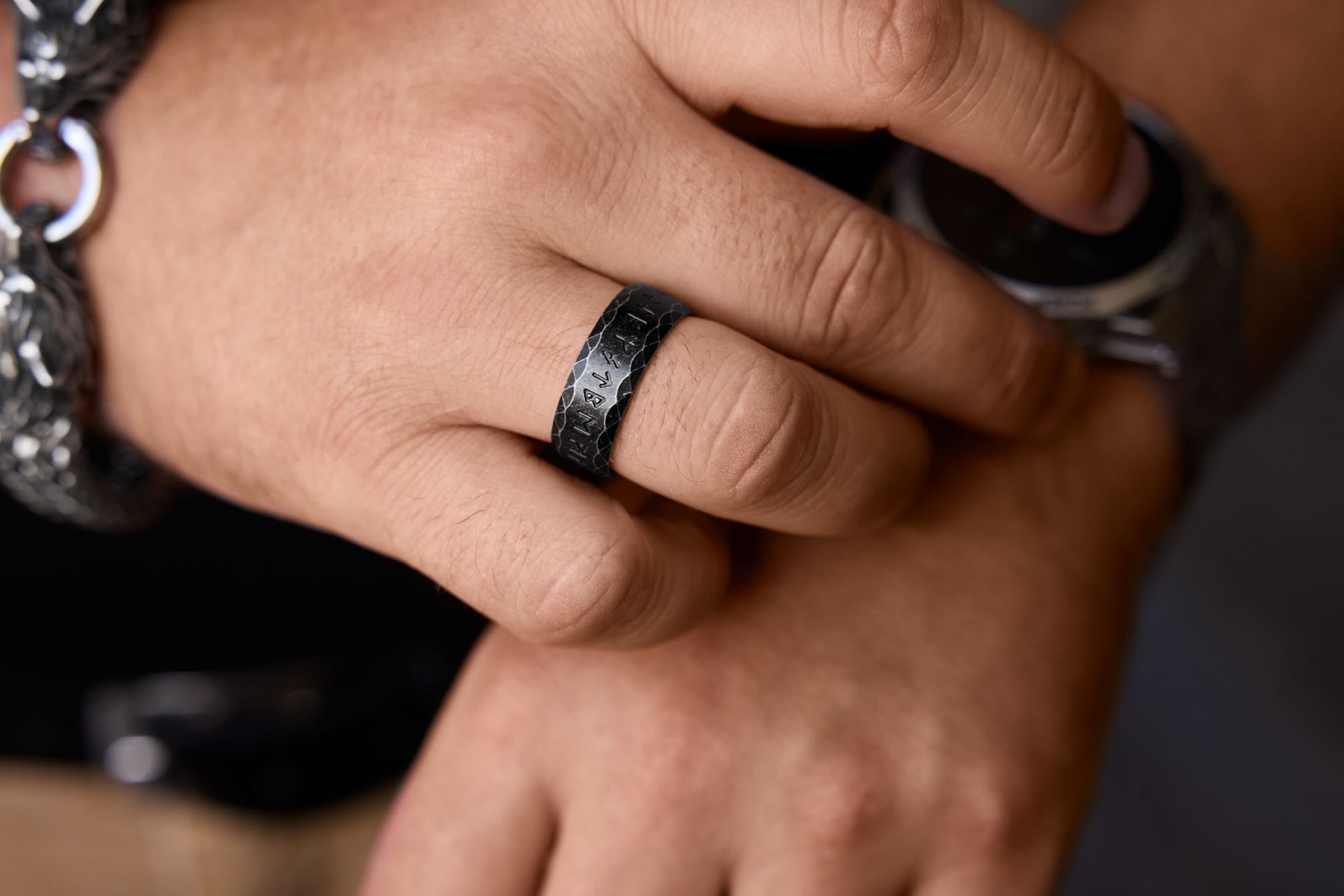 Close-up of a hand wearing the "Antique" variation stainless steel rune ring with engraved Elder Futhark symbols. The dark-toned ring is paired with a bold wolf-themed bracelet, adding a touch of Norse mythology-inspired style to the look.