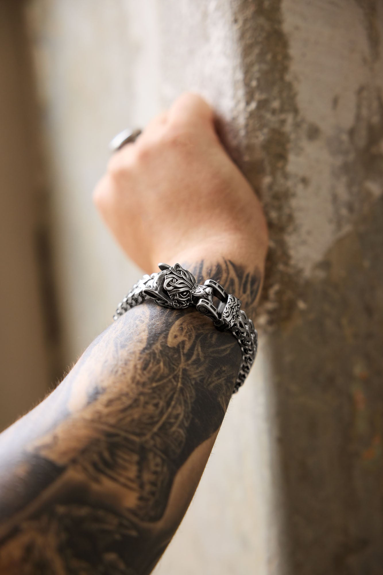 Close-up of a Fenrir bracelet with a detailed wolf head clasp, worn on a tattooed arm featuring a Fenrir-inspired design, leaning against a textured concrete wall. Available at Odin Trinkets.