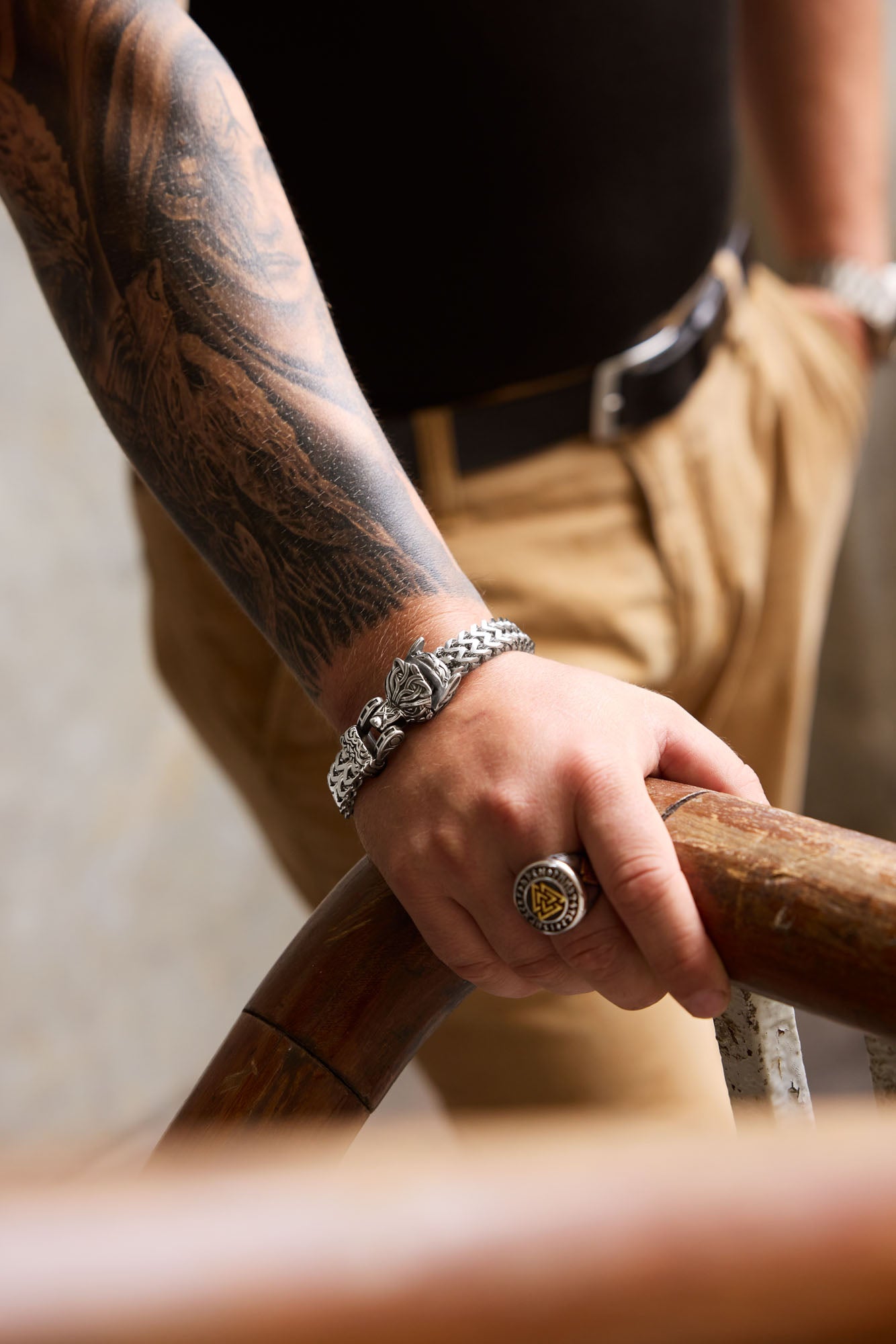 Man wearing a Fenrir bracelet with a wolf head clasp and a Valknut ring featuring a Nordic symbol, paired with a detailed tattoo sleeve, leaning on a wooden railing. Available at Odin Trinkets.