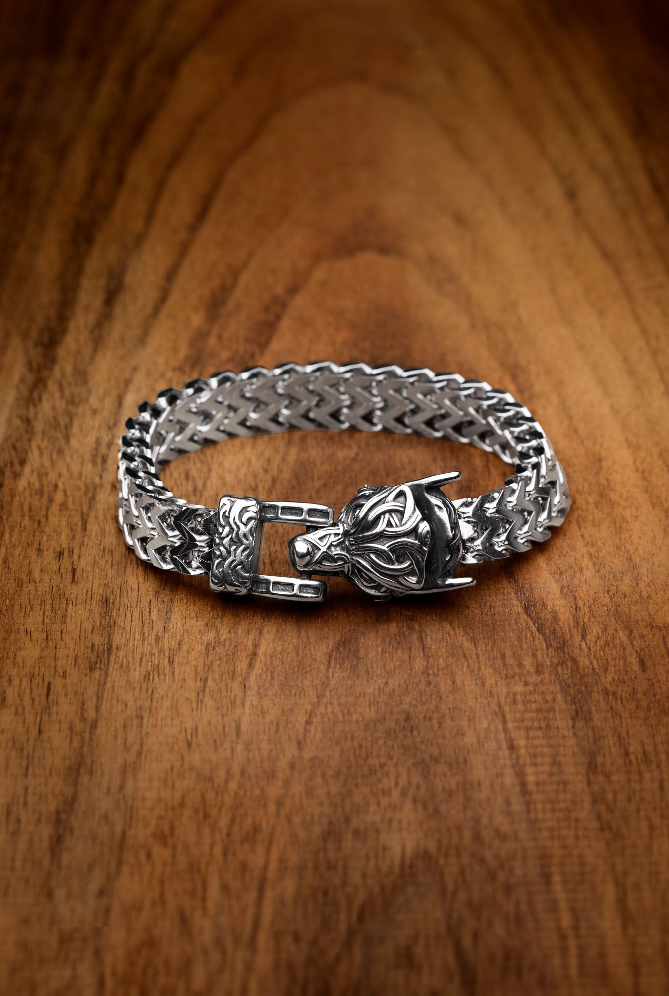 enrir bracelet featuring a silver wolf head clasp, inspired by the legendary wolf Fenrir from Norse mythology, with a woven chain design displayed on a wooden surface. Available at Odin Trinkets.