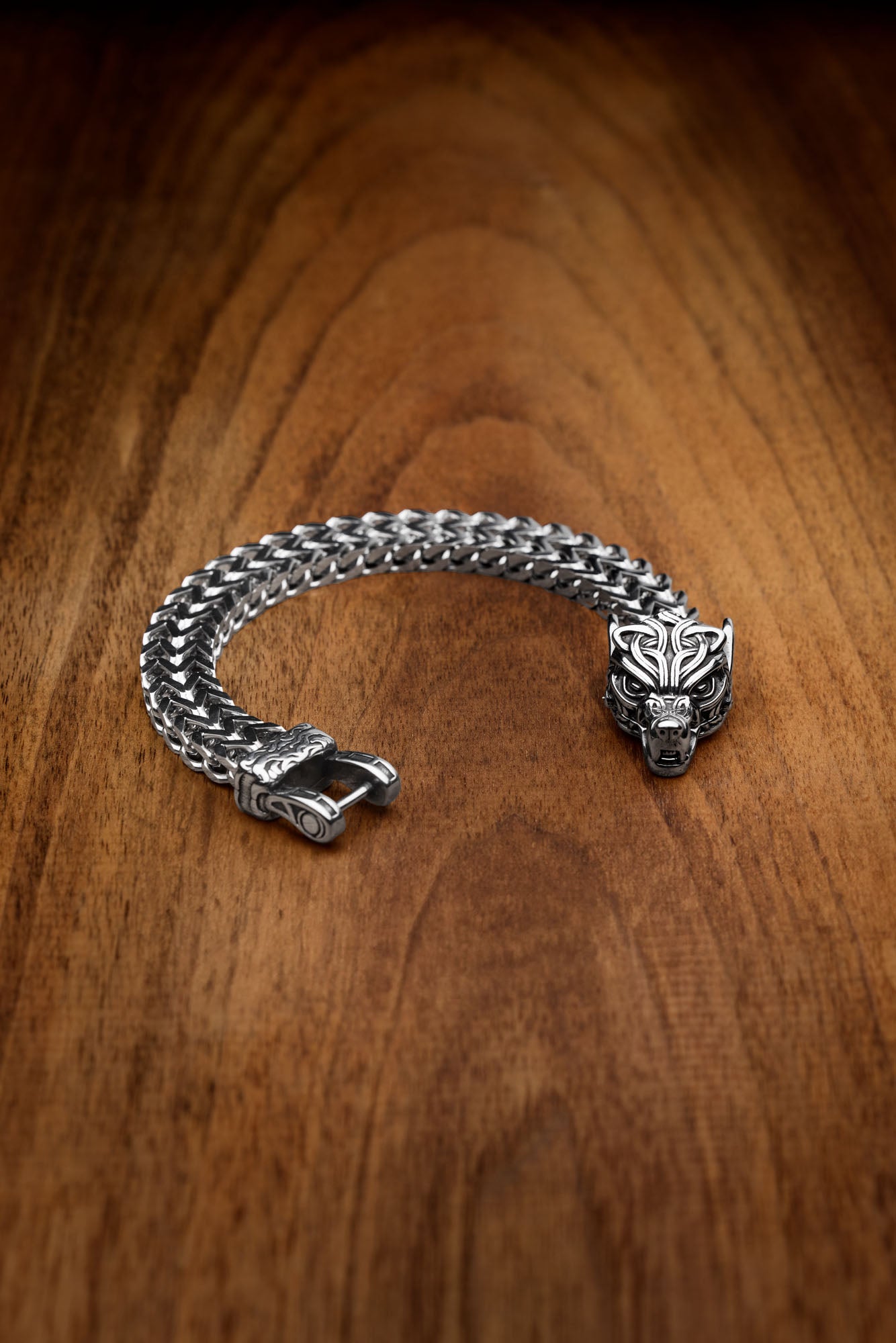 Fenrir bracelet with a silver wolf head clasp, inspired by the legendary wolf Fenrir from Norse mythology, featuring a woven chain design and displayed on a wooden surface. Available at Odin Trinkets.