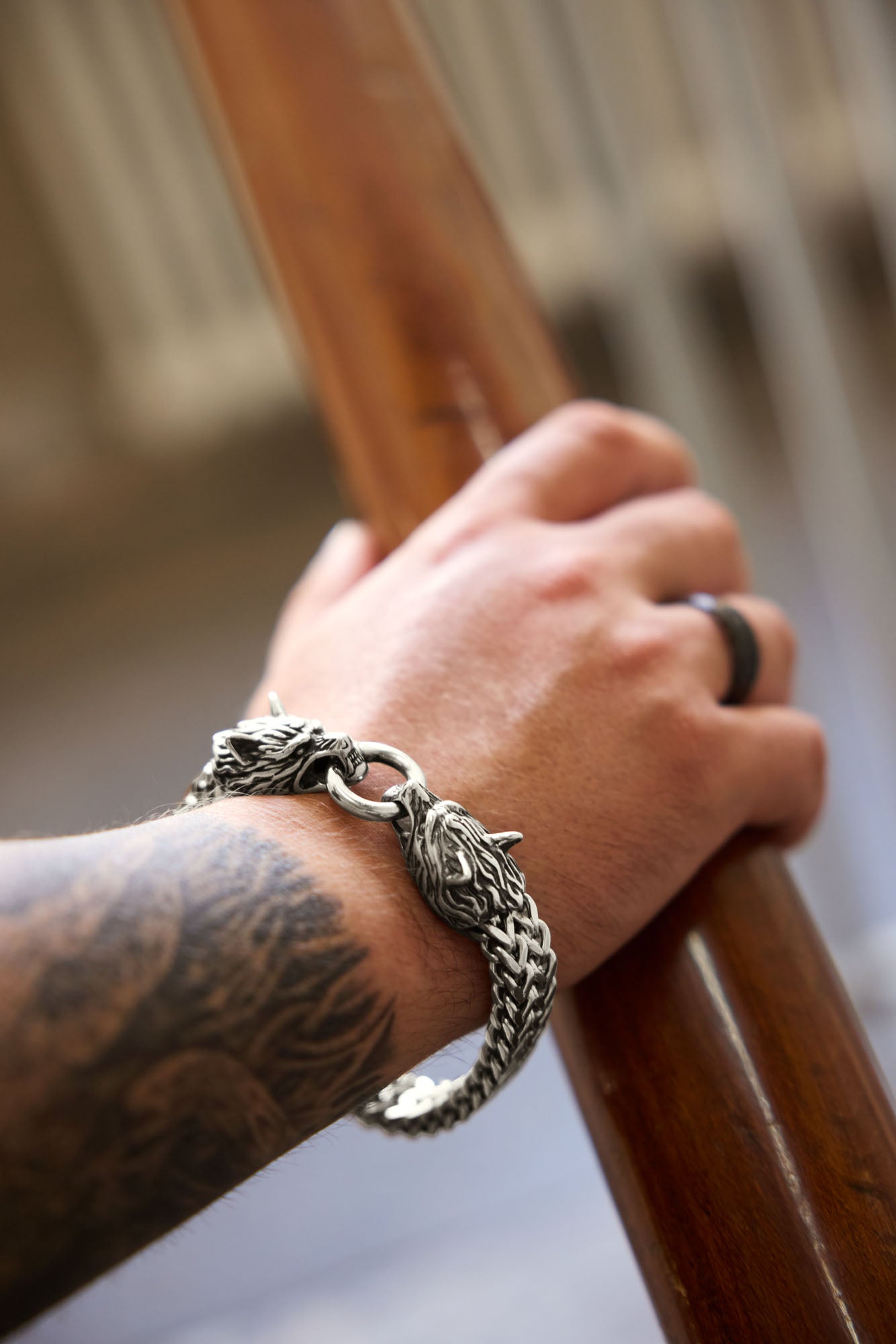 Close-up of a hand wearing the Geri and Freki bracelet with detailed wolf heads and an antique runic ring, paired with a tattooed arm, resting on a wooden railing. Available at Odin Trinkets.