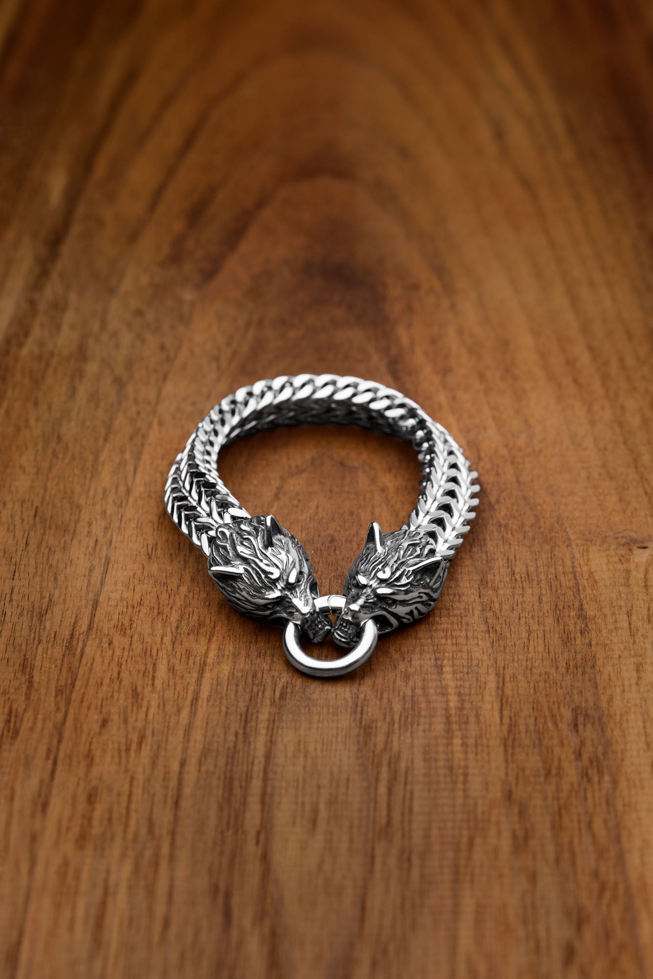 Geri and Freki bracelet featuring two intricately designed wolf heads clasping a metal ring, inspired by the legendary wolves of Norse mythology, displayed on a wooden surface. Available at Odin Trinkets.