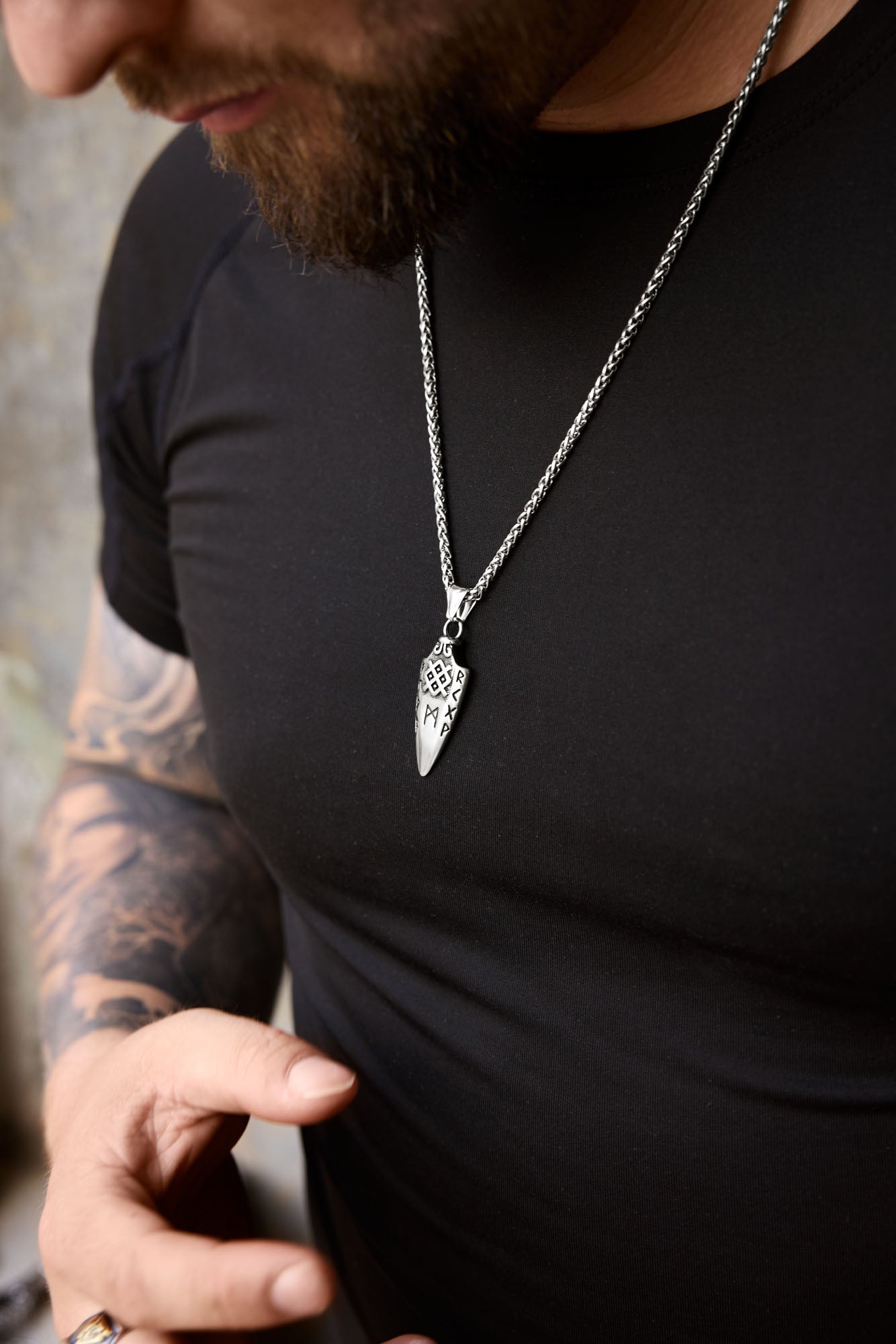 Close-up of a man wearing the Gungnir necklace with a spearhead pendant engraved with Nordic runes, paired with a tattooed arm and a black shirt, showcasing Norse mythology-inspired jewelry. Available at Odin Trinkets.