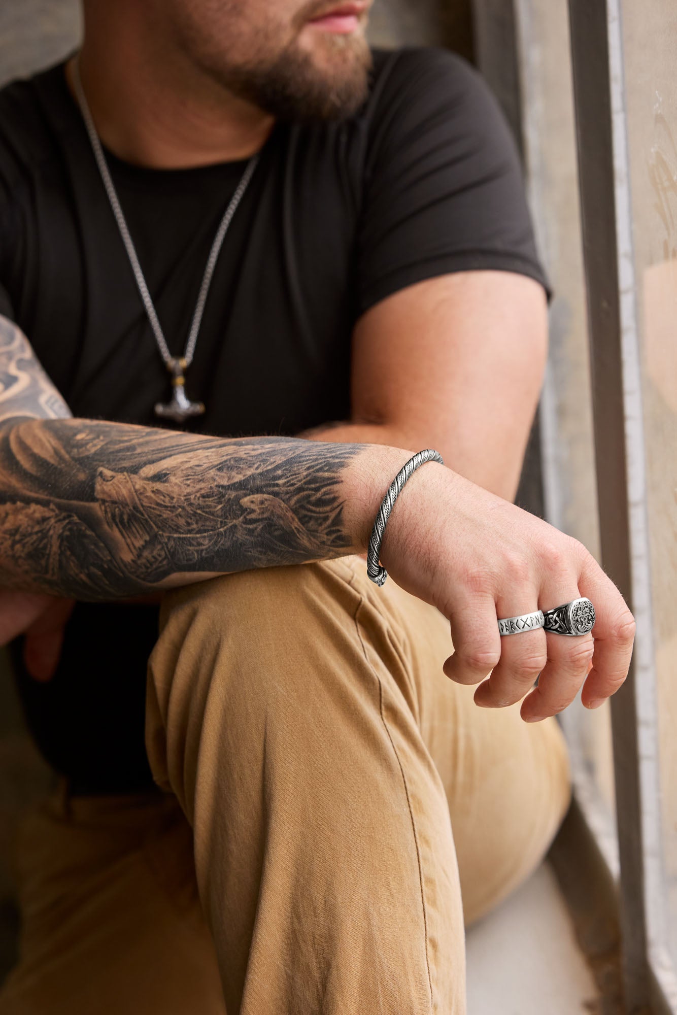 Man wearing the Mjölnir necklace with a Thor's Hammer pendant featuring intricate engravings and gold accents, paired with a twisted cuff bracelet, a silver rune ring showcasing the Elder Futhark alphabet, and a Yggdrasil ring symbolizing the Tree of Life, complemented by a tattooed arm and casual attire. Available at Odin Trinkets.