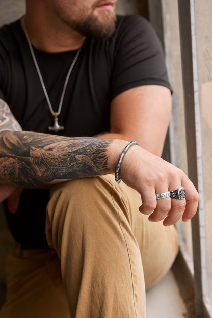 Man wearing the Mjölnir necklace with a Thor's Hammer pendant featuring intricate engravings and gold accents, paired with a twisted cuff bracelet, a silver rune ring showcasing the Elder Futhark alphabet, and a Yggdrasil ring symbolizing the Tree of Life, complemented by a tattooed arm and casual attire. Available at Odin Trinkets.