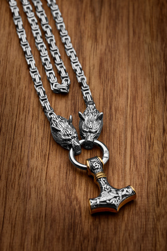 Mjölnir necklace with a Thor's Hammer pendant featuring intricate engravings and gold accents paired with a king chain design and wolf head clasps representing Geri and Freki the legendary wolves from Norse mythology displayed on a wooden surface Available at Odin Trinkets