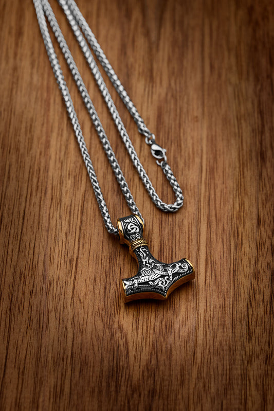 Mjölnir necklace featuring a detailed Thor's Hammer pendant with intricate engravings and gold accents, symbolizing strength and protection in Norse mythology, displayed on a wooden surface. Available at Odin Trinkets.