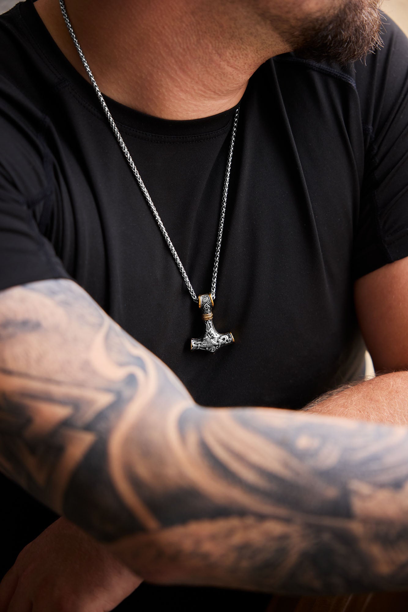 Man wearing the Mjölnir necklace with a Thor's Hammer pendant featuring intricate engravings and gold accents, paired with a tattooed arm and a black shirt, showcasing Norse mythology-inspired jewelry. Available at Odin Trinkets.