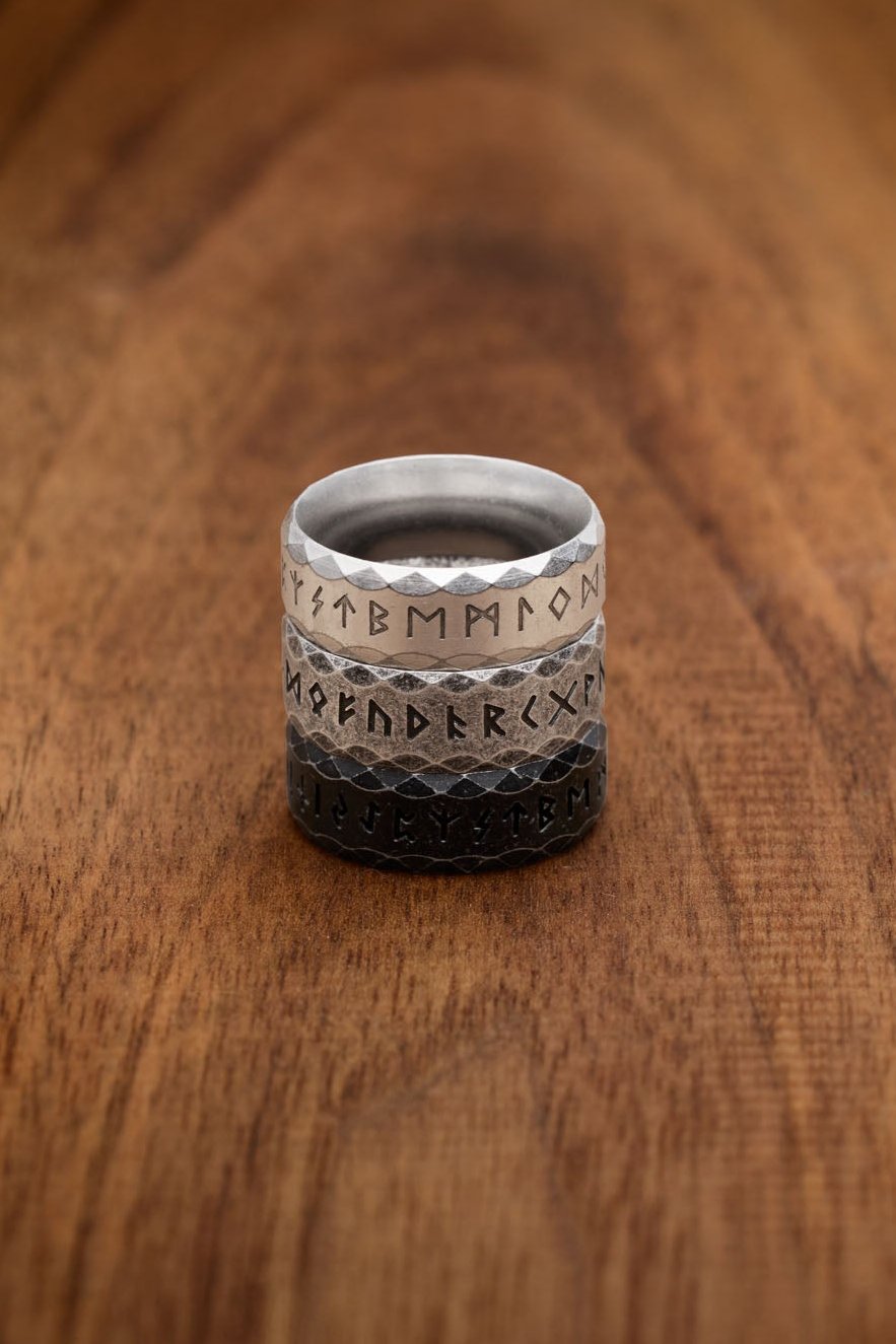  A stacked display of three stainless steel rune rings engraved with the complete Elder Futhark alphabet. The top ring in silver represents the "Silver" variation with a polished and reflective finish. The middle ring, in a weathered gray, is the "Retro" variation offering a rugged and aged look. The bottom ring features the "Antique" variation in black, with a dark and matte appearance, evoking ancient Norse designs.