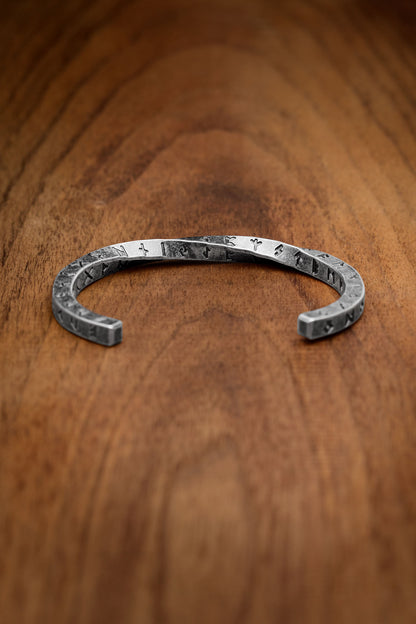 Twisted square design cuff bracelet engraved with the Elder Futhark alphabet, an ancient Nordic rune script, displayed on a wooden surface emphasizing its Viking-inspired aesthetic and craftsmanship Available at Odin Trinkets