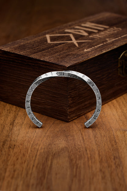 Twisted square design cuff bracelet engraved with Nordic runes symbolizing ancient Norse culture displayed in front of a wooden box with the Odin Trinkets logo showcasing Viking-inspired craftsmanship Available at Odin Trinkets