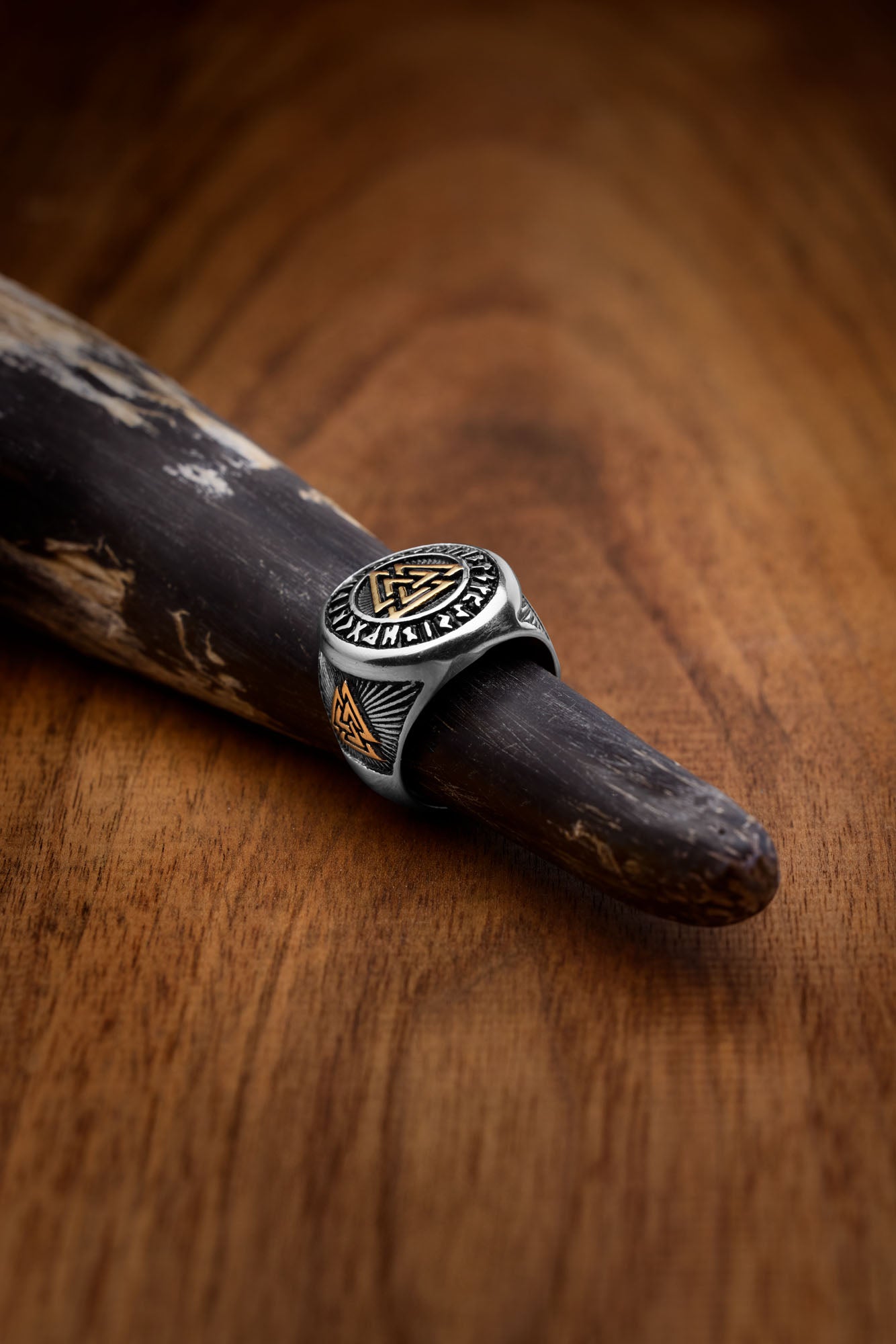 A stainless steel ring featuring the Valknut symbol in golden accents, surrounded by engraved Elder Futhark runes. The polished silver base contrasts with its intricate Norse design, resting elegantly on a rustic drinking horn.