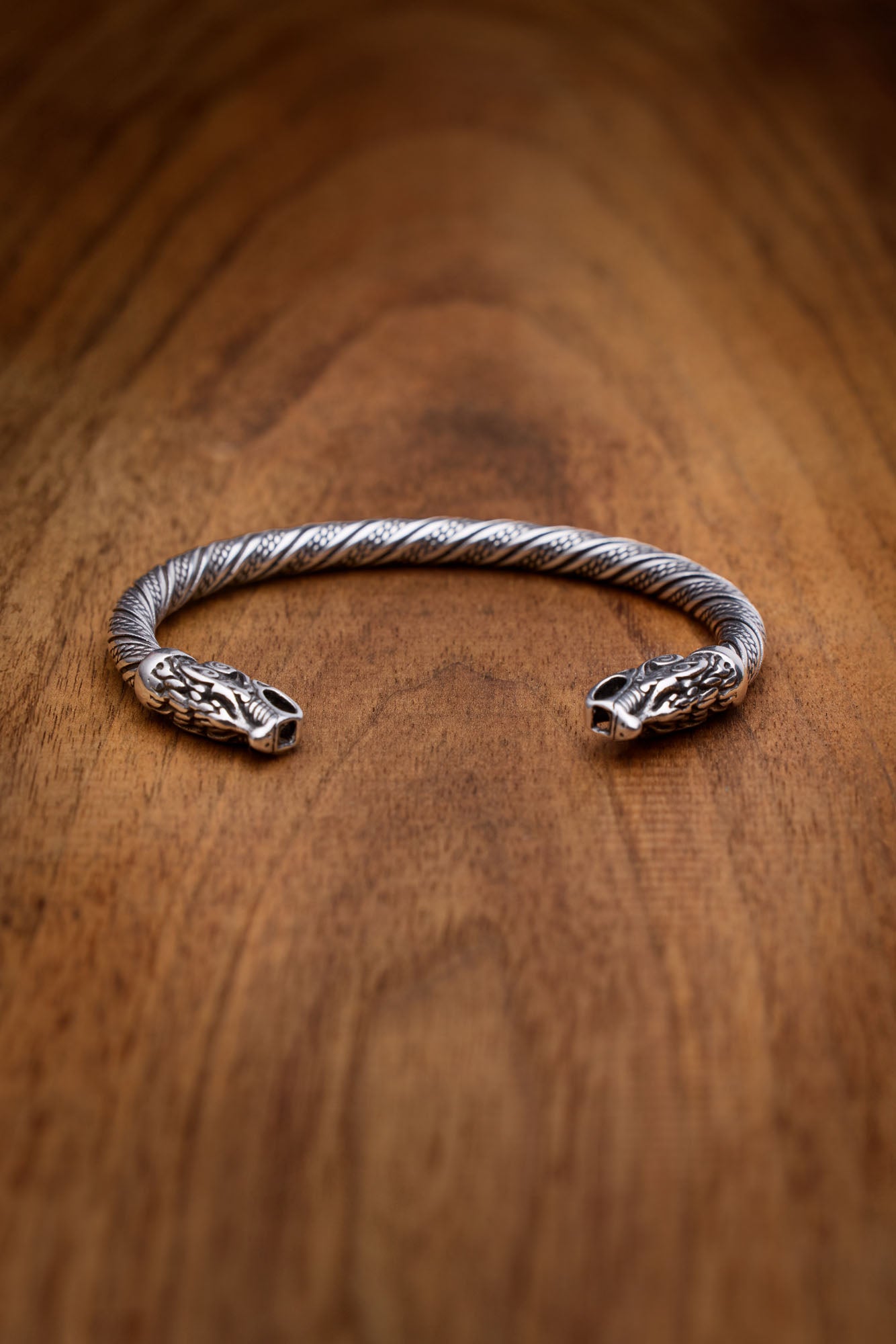 Stainless steel twisted cuff bracelet with intricately detailed wolf heads representing Geri and Freki, displayed against a wooden surface that emphasizes its Norse mythology-inspired design.